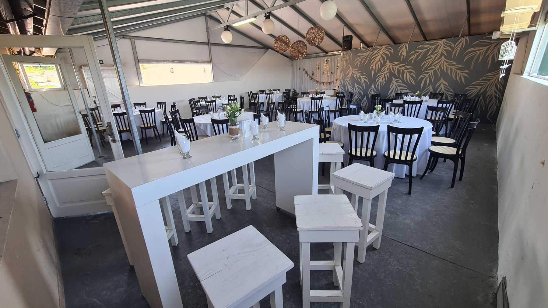 Patio techado con barra alta blanca, banquetas y mesas redondas con mural de hojas