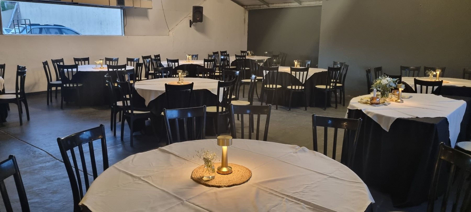 Salón armado con mesas redondas, manteles blancos y negros, y lámparas doradas